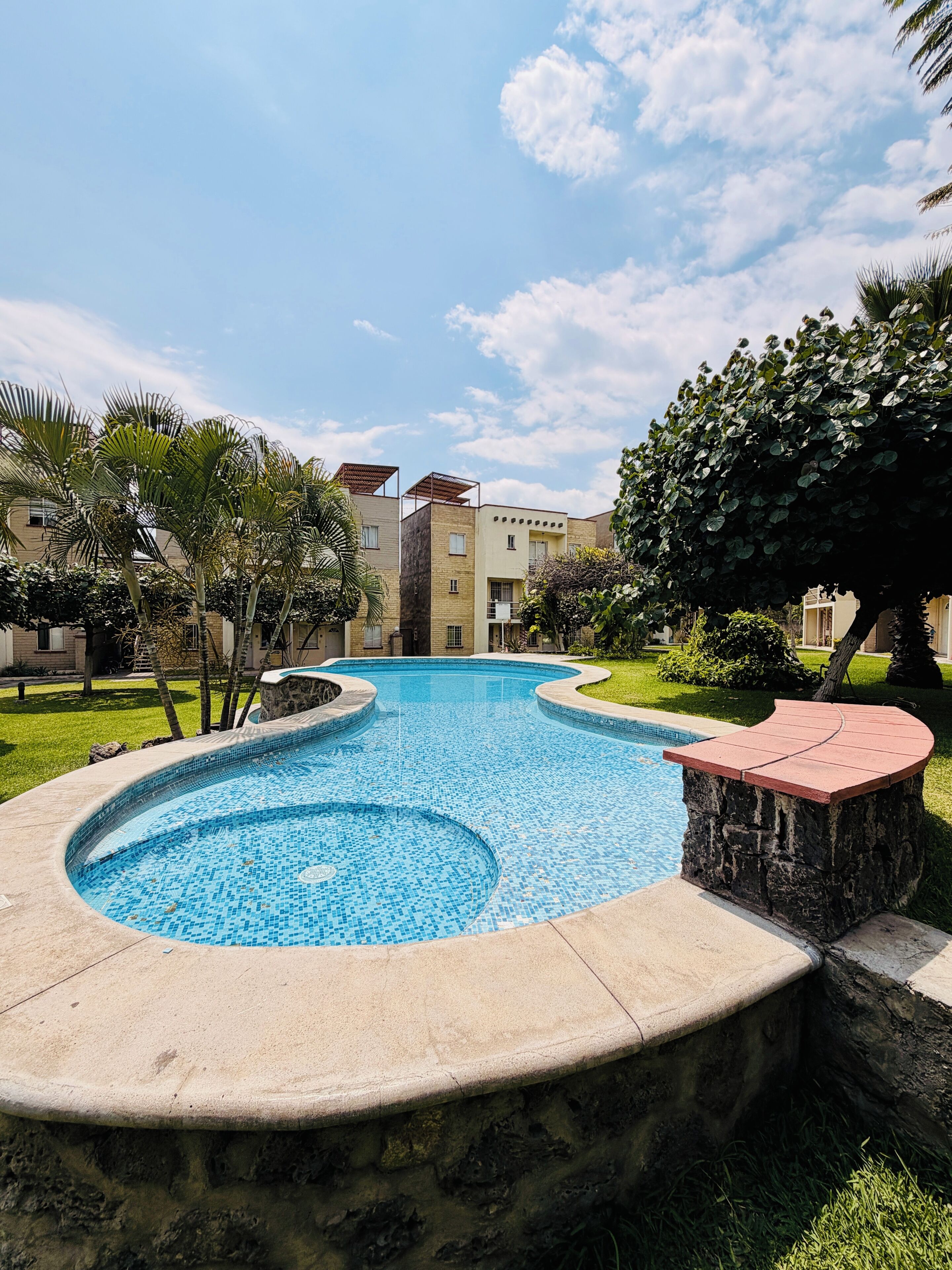 A swimming pool with a stone edge, surrounded by greenery and a building in the background.