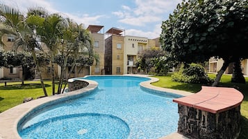 A swimming pool with a stone edge, surrounded by greenery and a building in the background.