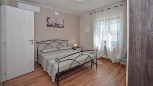 Ein Schlafzimmer mit einem Bett, einem Nachttisch, einer Wand-Klimaanlage, einem Fenster mit leichtem Vorhang und Holzboden.