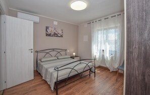 Ein Schlafzimmer mit einem Bett, einem Nachttisch, einer Wand-Klimaanlage, einem Fenster mit leichtem Vorhang und Holzboden.