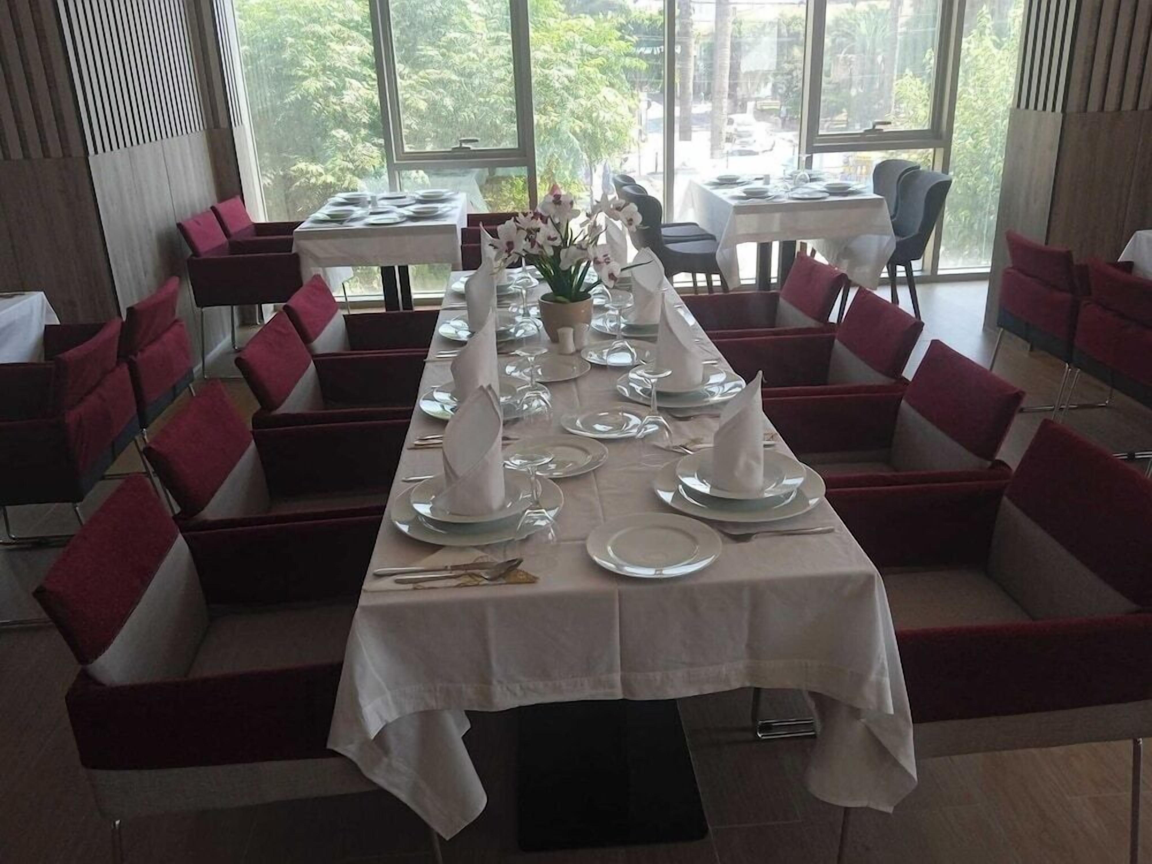 Un restaurant avec une longue table dressée pour un repas, des chaises rembourrées de couleur rouge et une vue dégagée sur l’extérieur grâce à de grandes fenêtres.