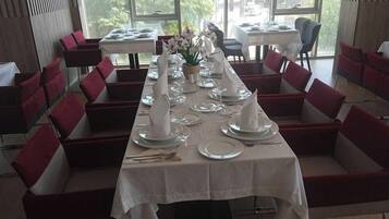 Un restaurant avec une longue table dressée pour un repas, des chaises rembourrées de couleur rouge et une vue dégagée sur l’extérieur grâce à de grandes fenêtres.