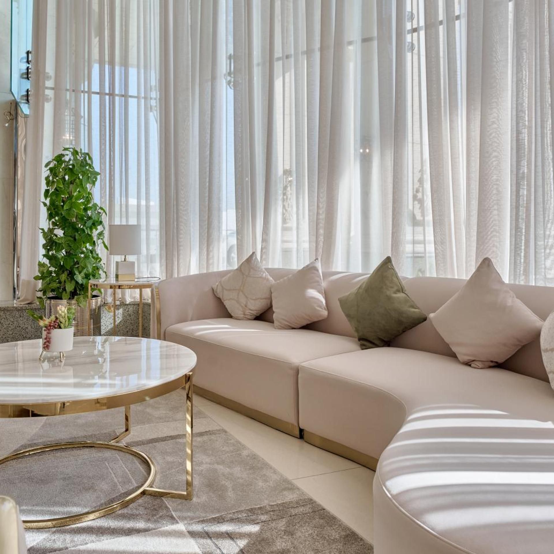 Una sala moderna con un sofá color beige, una mesa redonda de centro con patas doradas y cortinas traslúcidas.