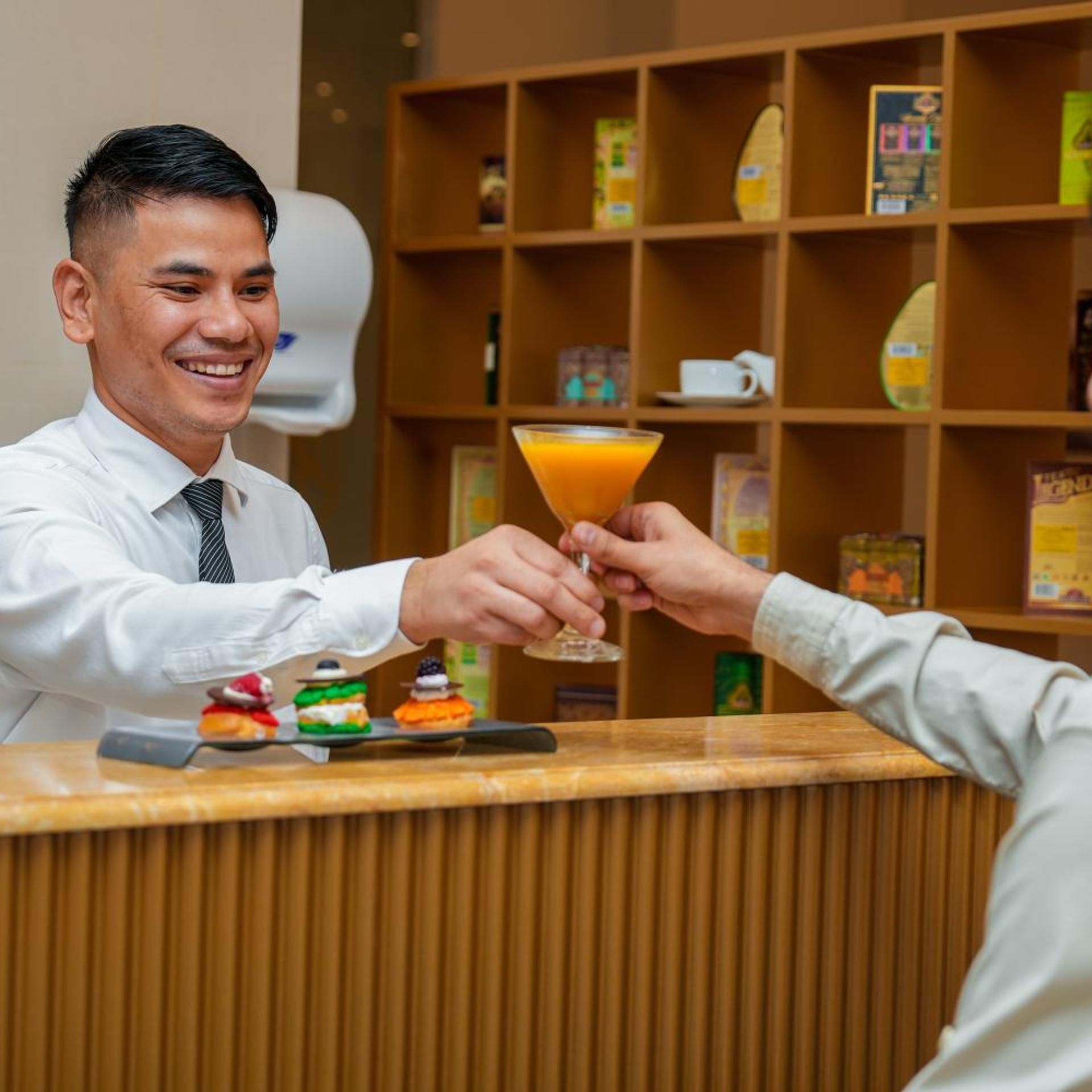 A person at a reception desk holding a martini glass, with a tray of desserts in front.