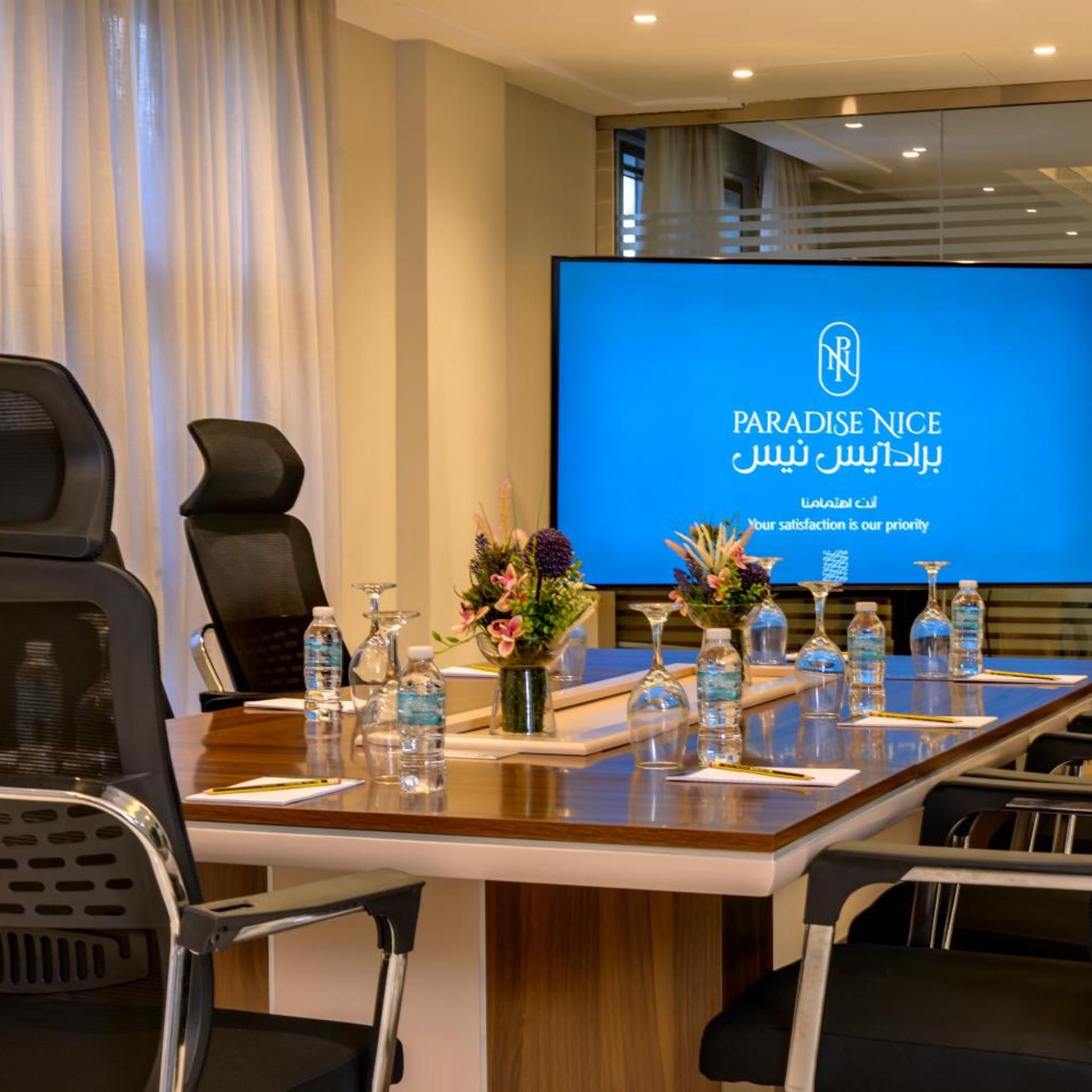 A conference room with a large screen displaying 'PARADISE NICE' and a logo, a long table with water bottles, vases with flowers, and black chairs.
