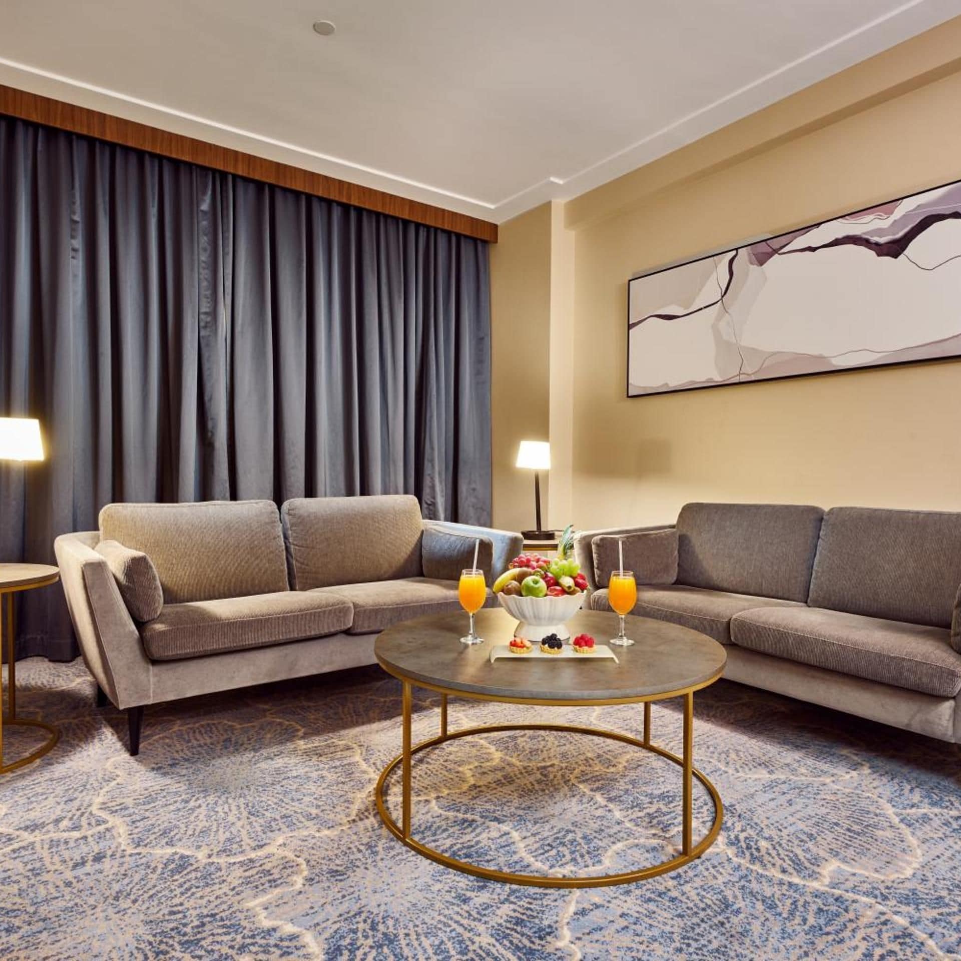 A hotel room with a sofa set, a round coffee table with fruit and drinks, a large abstract painting on the wall, and a patterned carpet.