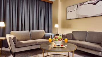 A hotel room with a sofa set, a round coffee table with fruit and drinks, a large abstract painting on the wall, and a patterned carpet.