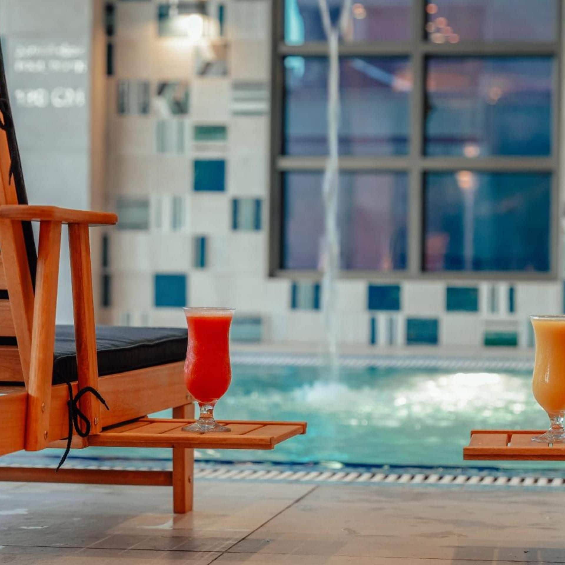 Un área junto a la piscina con una silla de descanso y dos vasos de bebidas sobre una mesa de madera.