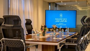 Una sala de conferencias con una pantalla grande mostrando 'PARADISE NICE' y texto en árabe, una mesa larga con sillas y botellas de agua.