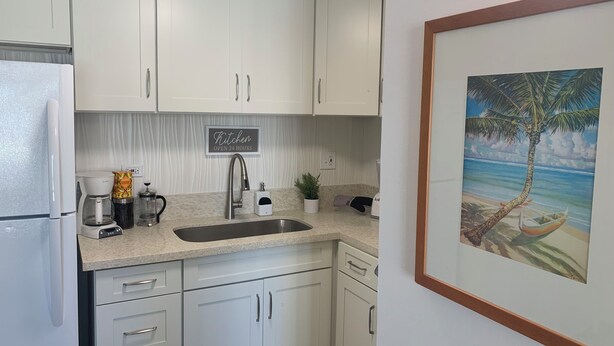Uma cozinha com armários brancos, uma pia de aço inoxidável, uma cafeteira e um quadro com uma paisagem de praia.