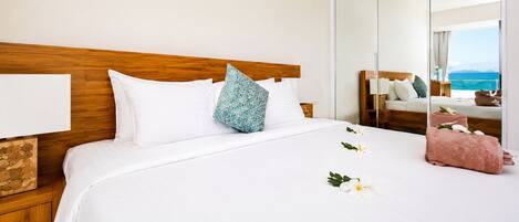 A hotel room with a wooden headboard, white bedding, a decorative pillow, a bedside table with a lamp, and a view of the sea.