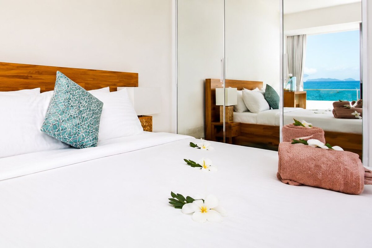 A hotel room with a wooden headboard, white bedding, a decorative pillow, a bedside table with a lamp, and a view of the sea.