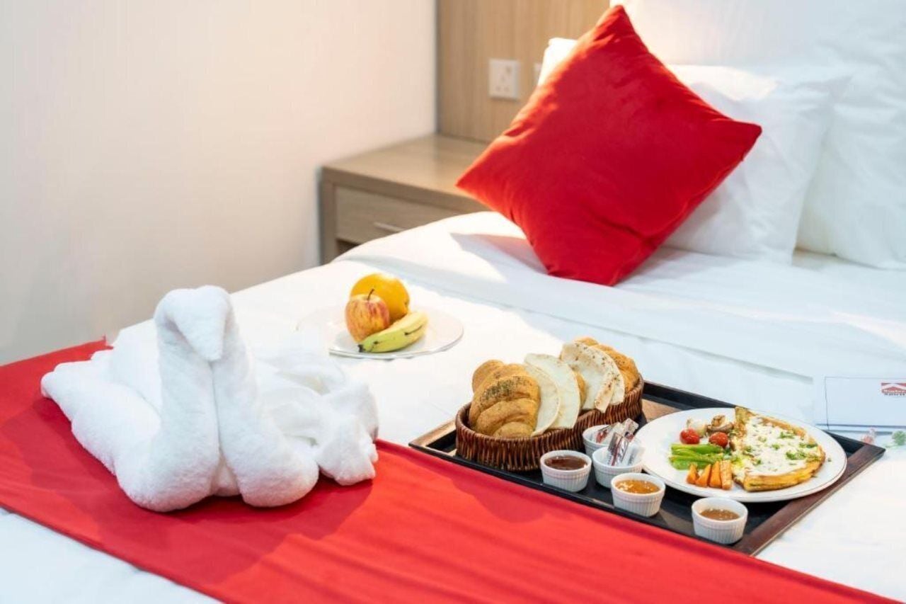 Een hotelkamer met een net opgemaakt bed, een dienblad met ontbijtartikelen, waaronder croissants, een bord pizza en een fruitschaal.