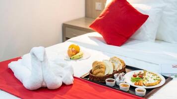 Een hotelkamer met een net opgemaakt bed, een dienblad met ontbijtartikelen, waaronder croissants, een bord pizza en een fruitschaal.