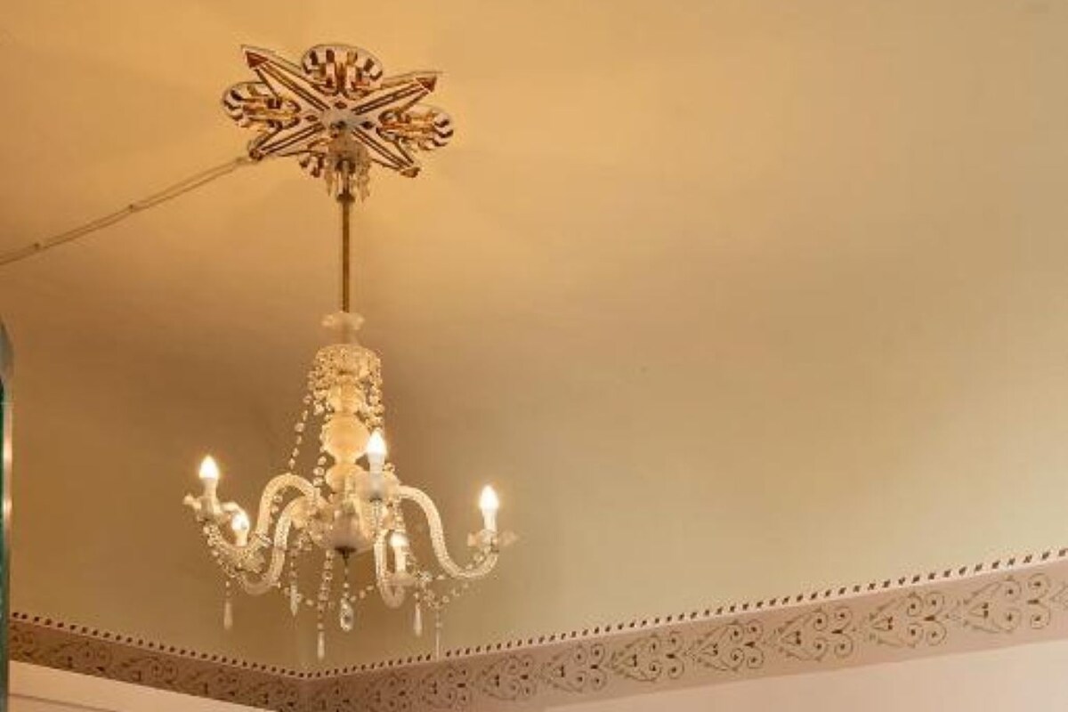 A bedroom with a large bed, a chandelier, a painting on the wall, and a bathroom visible in the background.