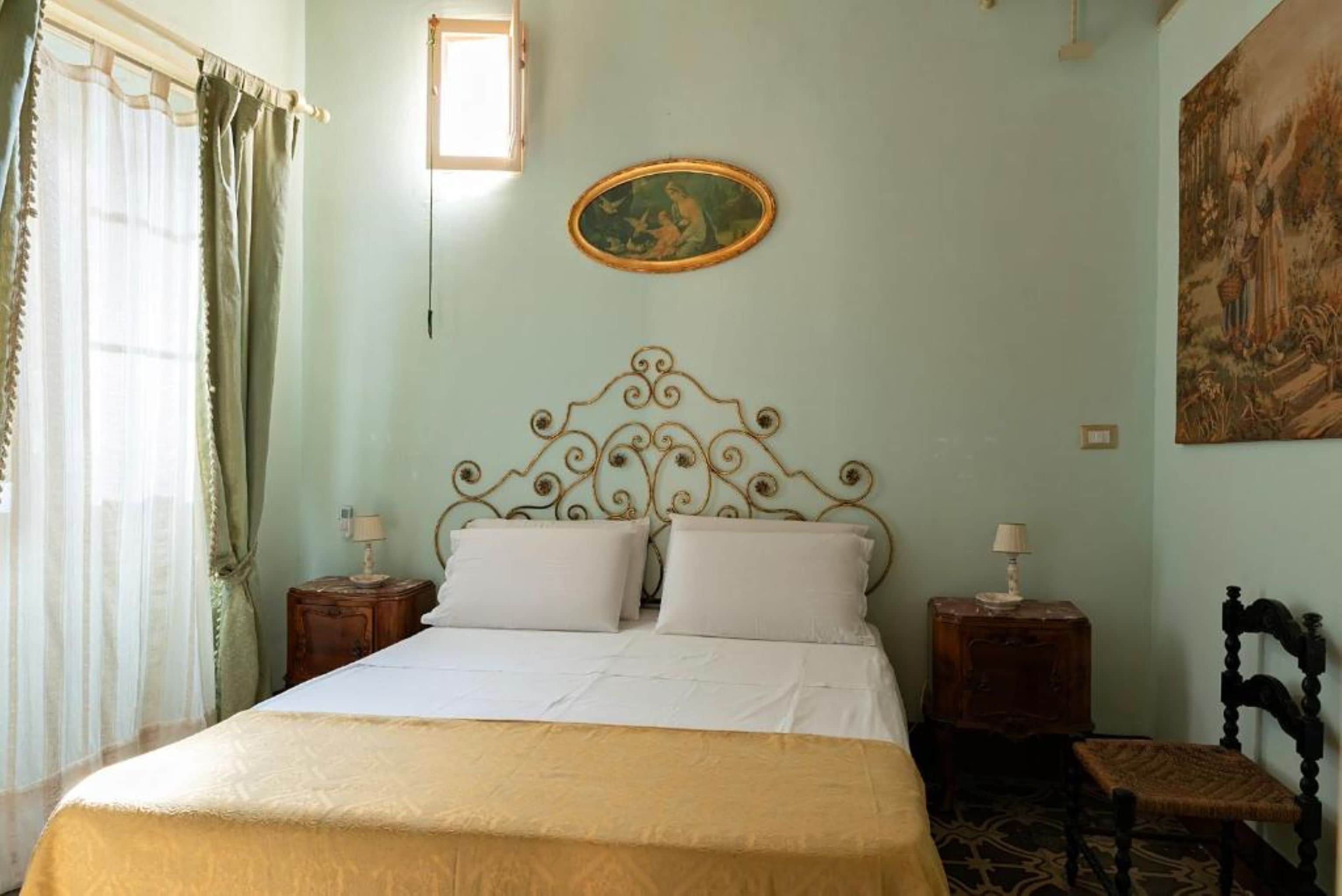 Une chambre à coucher avec un lit, deux tables de chevet, une chaise, un tableau accroché au mur et une fenêtre avec des rideaux.