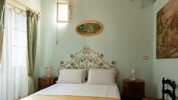 A bedroom with a bed, two bedside tables, a chair, a painting on the wall, and a window with curtains. - Roccamelia Experience (Mazara del Vallo)