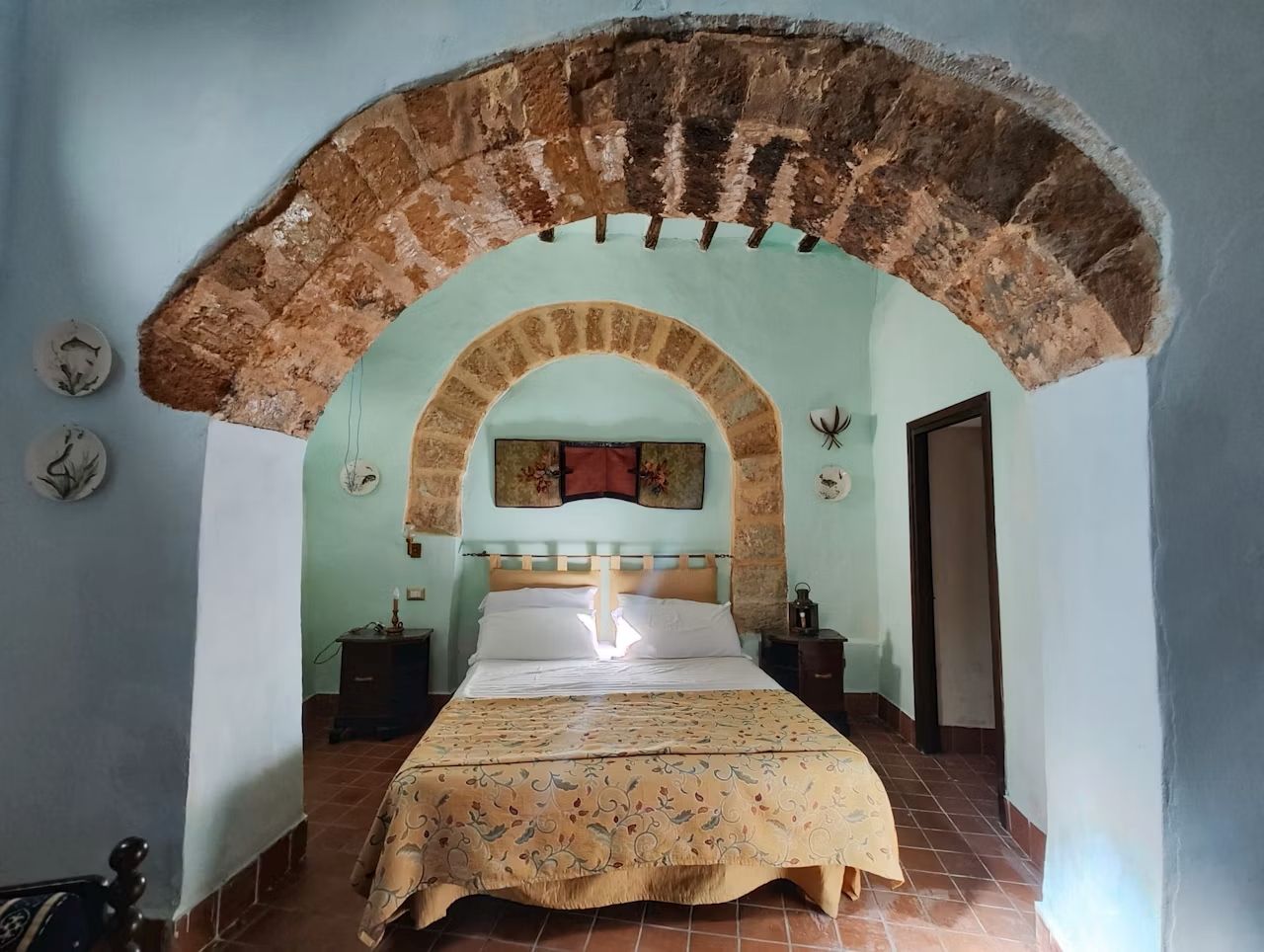 A bedroom with a bed centered under a stone archway, a nightstand, a wall clock, and a door to the right.