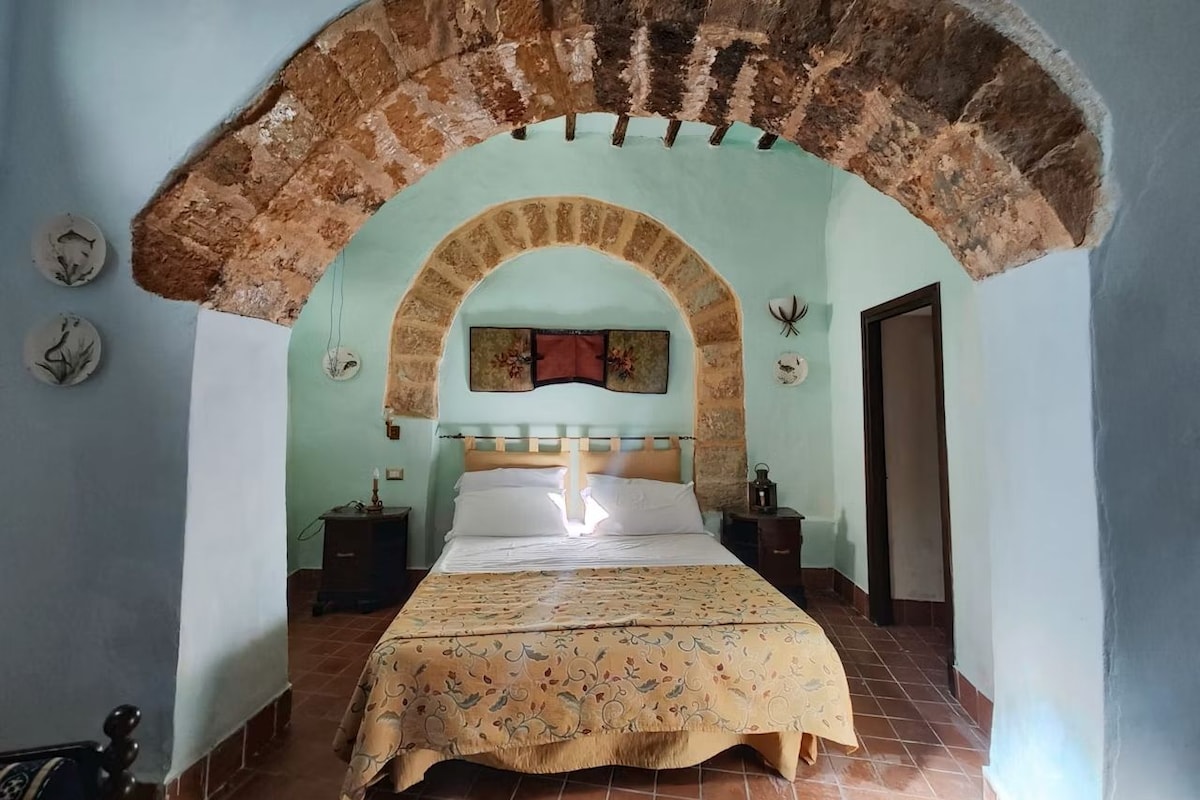 A bedroom with a bed centered under a stone archway, a nightstand, a wall clock, and a door to the right.