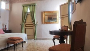 A hotel room with a bed, a desk, a chair, a mirror, and a window with green curtains. - Roccamelia Experience (Mazara del Vallo)