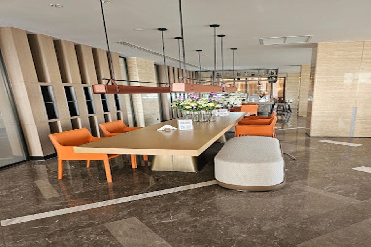 Un hall d’hôtel moderne avec une longue table rectangulaire, des chaises orange, un pouf blanc et un vase de fleurs.