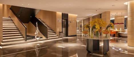 A modern lobby with a central staircase, a reception desk, and a decorative vase with flowers.