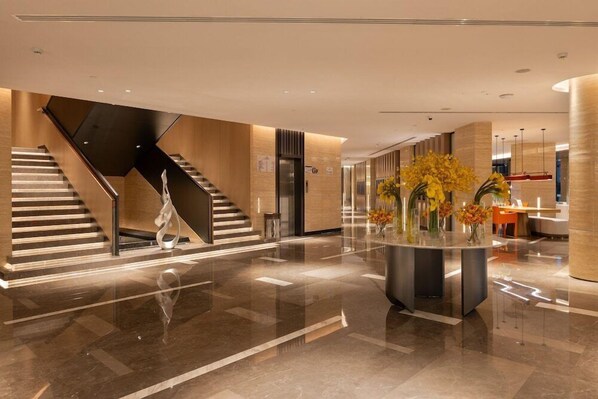 A modern lobby with a central staircase, a reception desk, and a decorative vase with flowers.