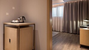 Habitación de hotel con un armario de madera, una mesa con hervidor, un escritorio con lámpara y vistas al exterior a través de cortinas.