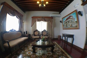 Una habitación con sofá, dos sillones, una mesa de centro, alfombra estampada, candelabro, un cuadro y aire acondicionado instalado en la pared.