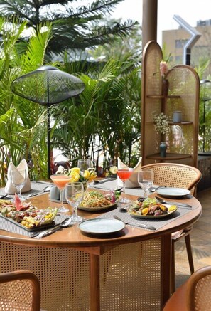 An outdoor dining area with round tables, wicker chairs, and a variety of dishes and drinks. - Urbane By Treat, Daman (Daman)