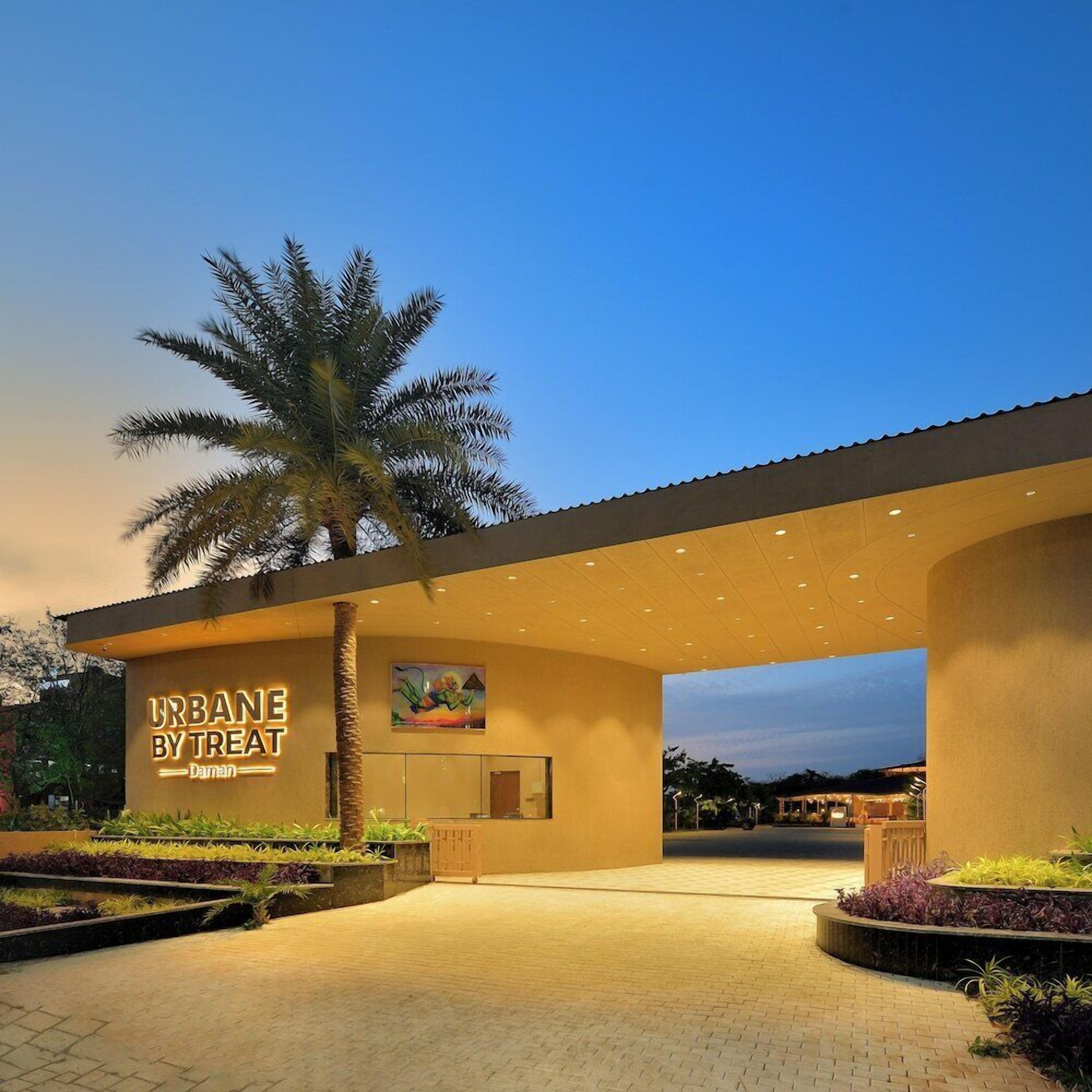 A modern building with a sign reading 'URBANE BY TREAT Daman' and a palm tree in front.