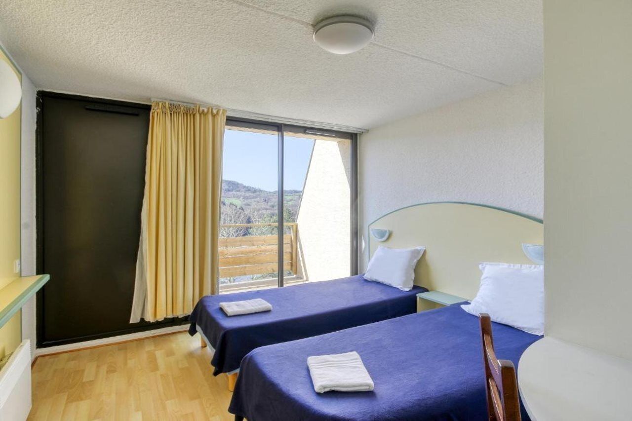 A room with two beds, a window with curtains, a wooden floor, and a view of the outdoors.