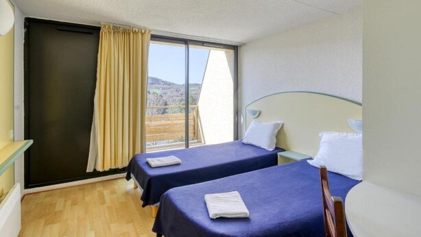Ein Zimmer mit zwei Betten, einem Vorhangfenster, Holzboden und Blick ins Freie.