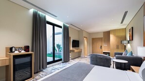 Ein modernes Hotelzimmer mit einem großen Bett, einer Sitzecke mit Sofa und Sesseln, einem kleinen Fernseher, einer Kaffeemaschine und Blick ins Freie durch eine Schiebetür.