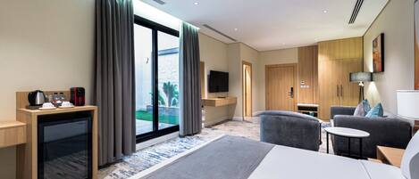 A modern hotel room with a large bed, a seating area with a sofa and armchairs, a small TV, a coffee machine, and a view of the outdoors through a sliding door.