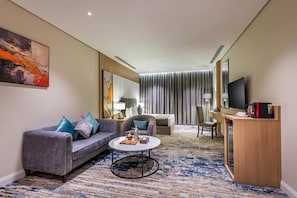 A modern hotel room with a grey sofa, a round coffee table, a desk with a flat-screen TV, and a large window with curtains. - Rhenium Hotel (Riyadh)