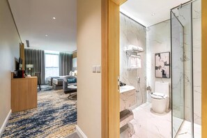 A hotel room with a marble bathroom, a sitting area with a sofa and coffee table, and a view of the city through large windows. - Rhenium Hotel (Riyadh)