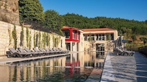 Una piscina con sillas de descanso, un edificio rojo y una ladera boscosa al fondo.