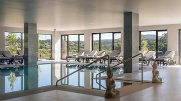 A modern indoor swimming pool with clear glass walls, lounge chairs, and a Buddha statue.