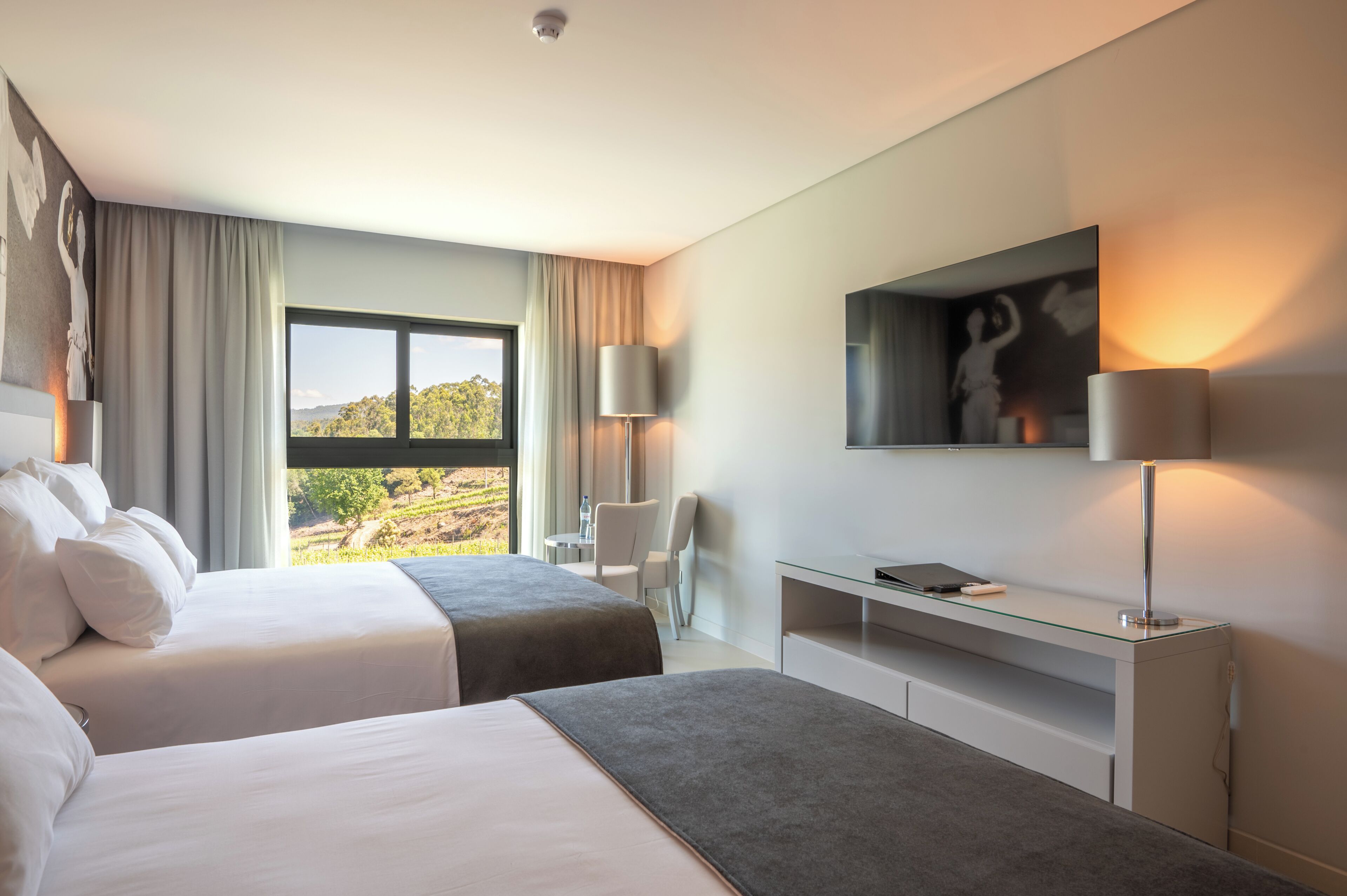 A hotel room with two beds, a desk, a chair, a TV, and a large window offering a view of the outdoors.