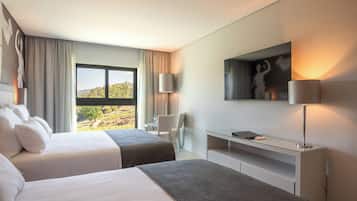 A hotel room with two beds, a desk, a chair, a TV, and a large window offering a view of the outdoors.