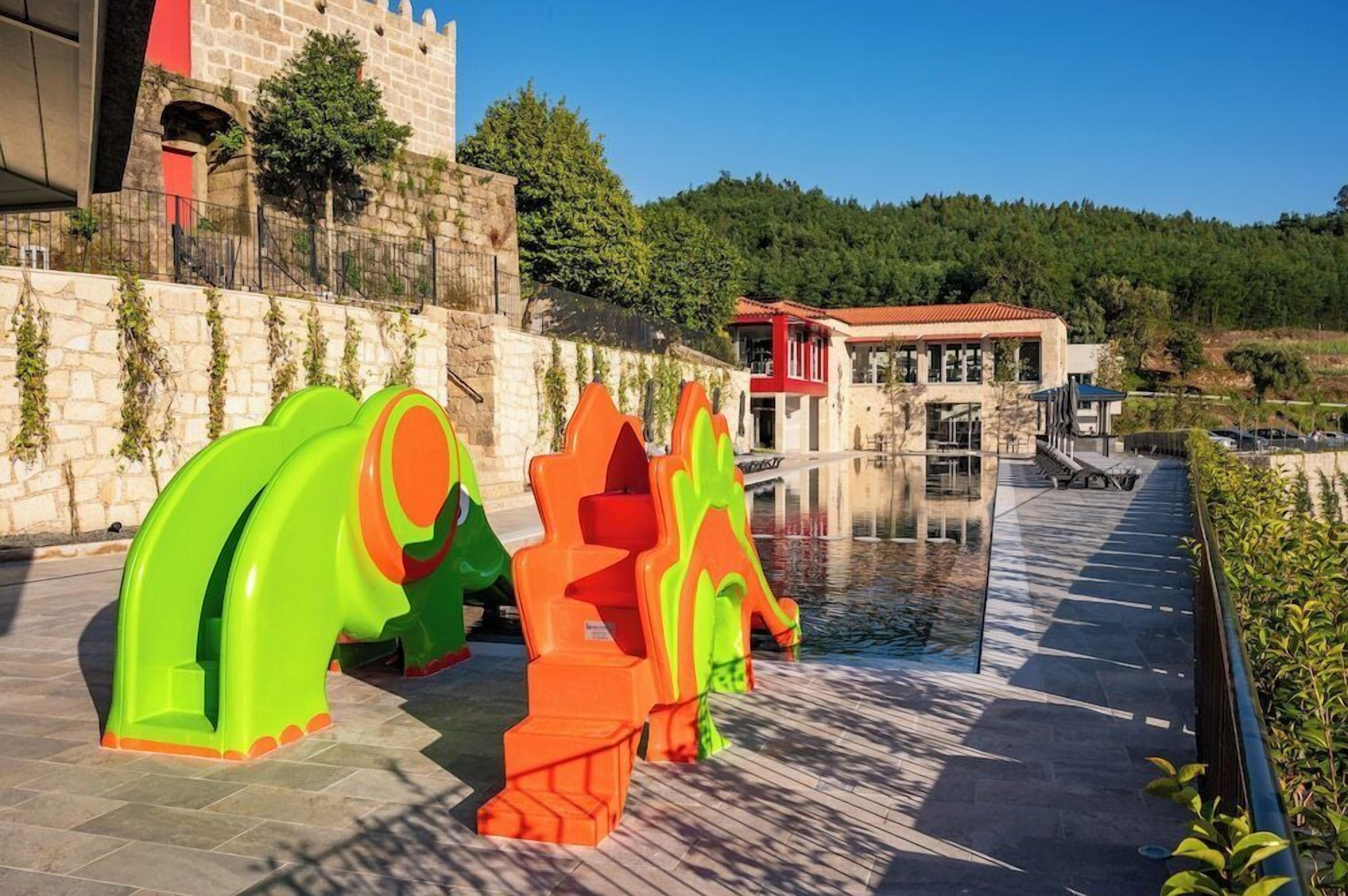 A waterfront area with a stone wall, a red-roofed building, and colorful abstract sculptures.