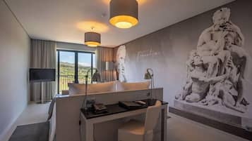 A modern hotel room with a desk, a large window, a flat-screen TV, and a wall-mounted sculpture.
