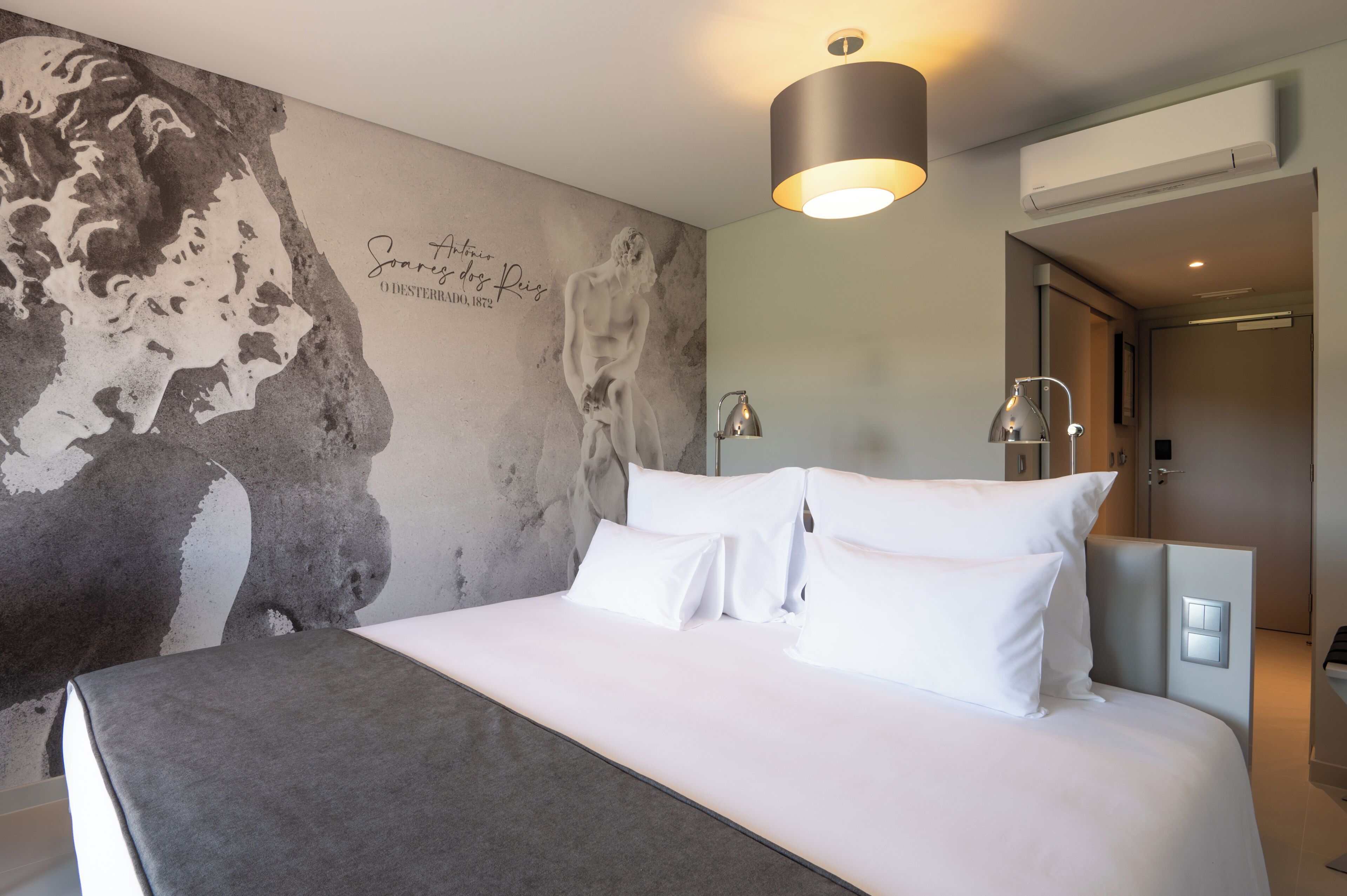 A modern hotel room with a large bed, a wall mural, and a ceiling light fixture.