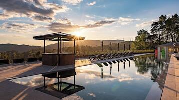 A pool with lounge chairs and a pergola, surrounded by trees and hills.