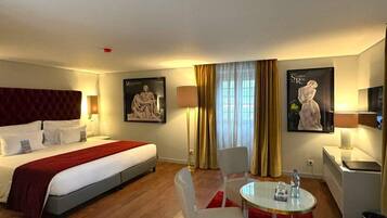 Una habitación de hotel con cama, escritorio, dos sillas, una mesa redonda y obras de arte en las paredes.