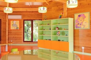 A room with wooden walls, a glass table, orange and green shelves with toys, and a large window. - Vila Gale Collection Ponte de Lima Vineyards (Ponte de Lima)