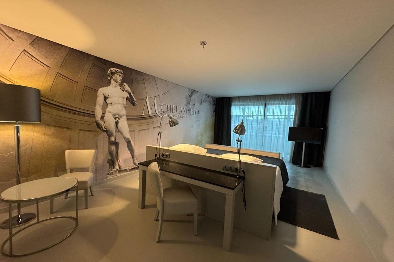 A hotel room with a bed, a desk, a chair, a lamp, and a large wall mural.