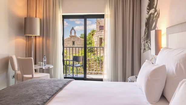 Habitación de hotel con una cama, una silla, una mesita con una botella y vista a un edificio de piedra a través de la puerta del balcón.