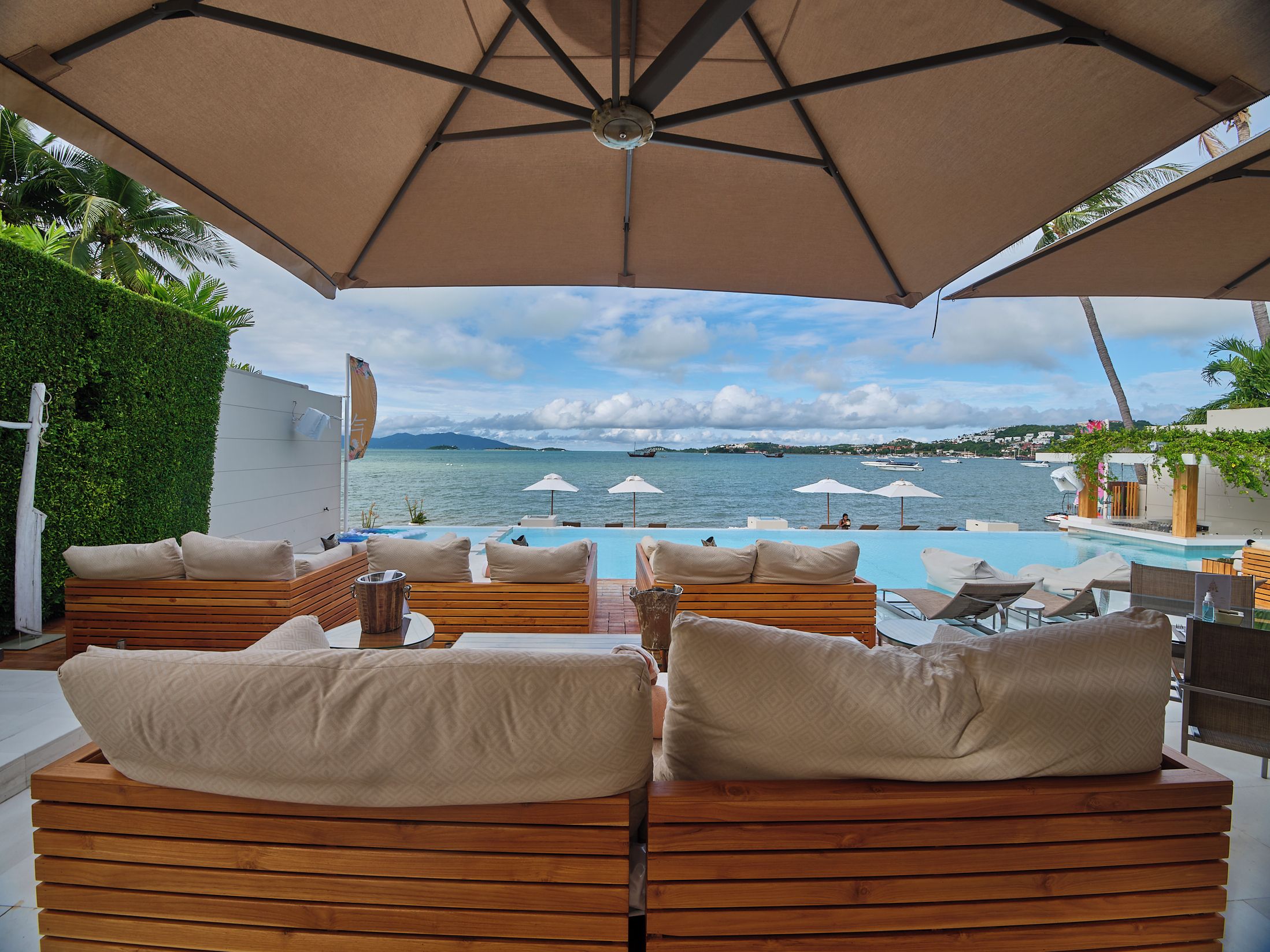 A poolside lounge area with wooden seating and umbrellas, overlooking a body of water with boats.